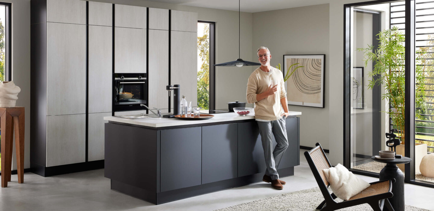 Moderne Burger Küche bei H&L Küchenstudio in Emlichheim mit Kochinsel in dunklem Design, hellen Hochschränken und einer Person im lichtdurchfluteten Raum mit großen Fensterflächen